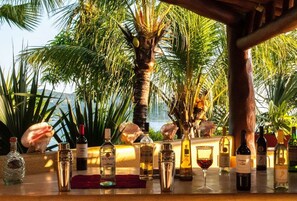 A bar area with various bottles and a glass, surrounded by palm trees and a view of the ocean. - Villa Encantada at Pacific Vacation | StayPV Oceanfront Luxury with Staff (Zihuatanejo)