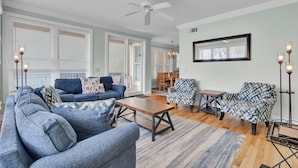 A living room with a sofa, armchairs, a coffee table, and a dining table. There is a ceiling fan, large windows with blinds, and a chandelier.