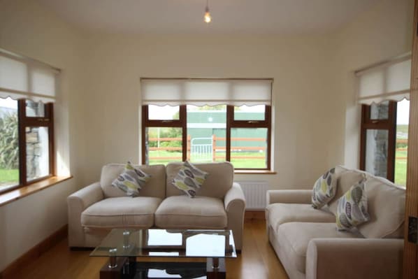A living room with two sofas, a glass coffee table, and large windows with blinds.