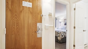 A hotel room with a wooden door, a bedside table, a lamp, a bed with floral bedding, and a small window with curtains. - Ski-In Whiffletree F5 Condo with King Bed, Weekend Shuttle & Balcony (Killington)