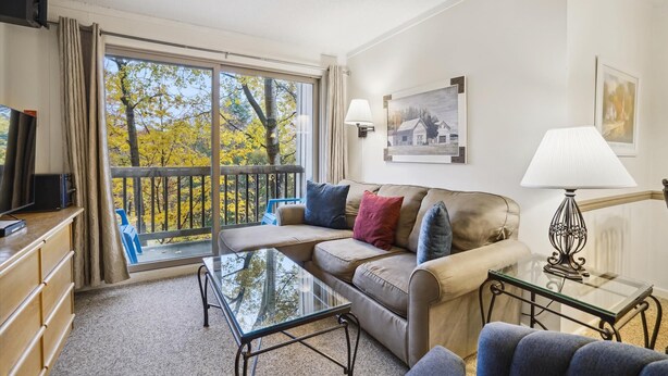 Een woonkamer met een bank, een glazen salontafel, een televisie en een balkon met herfstbladeren.