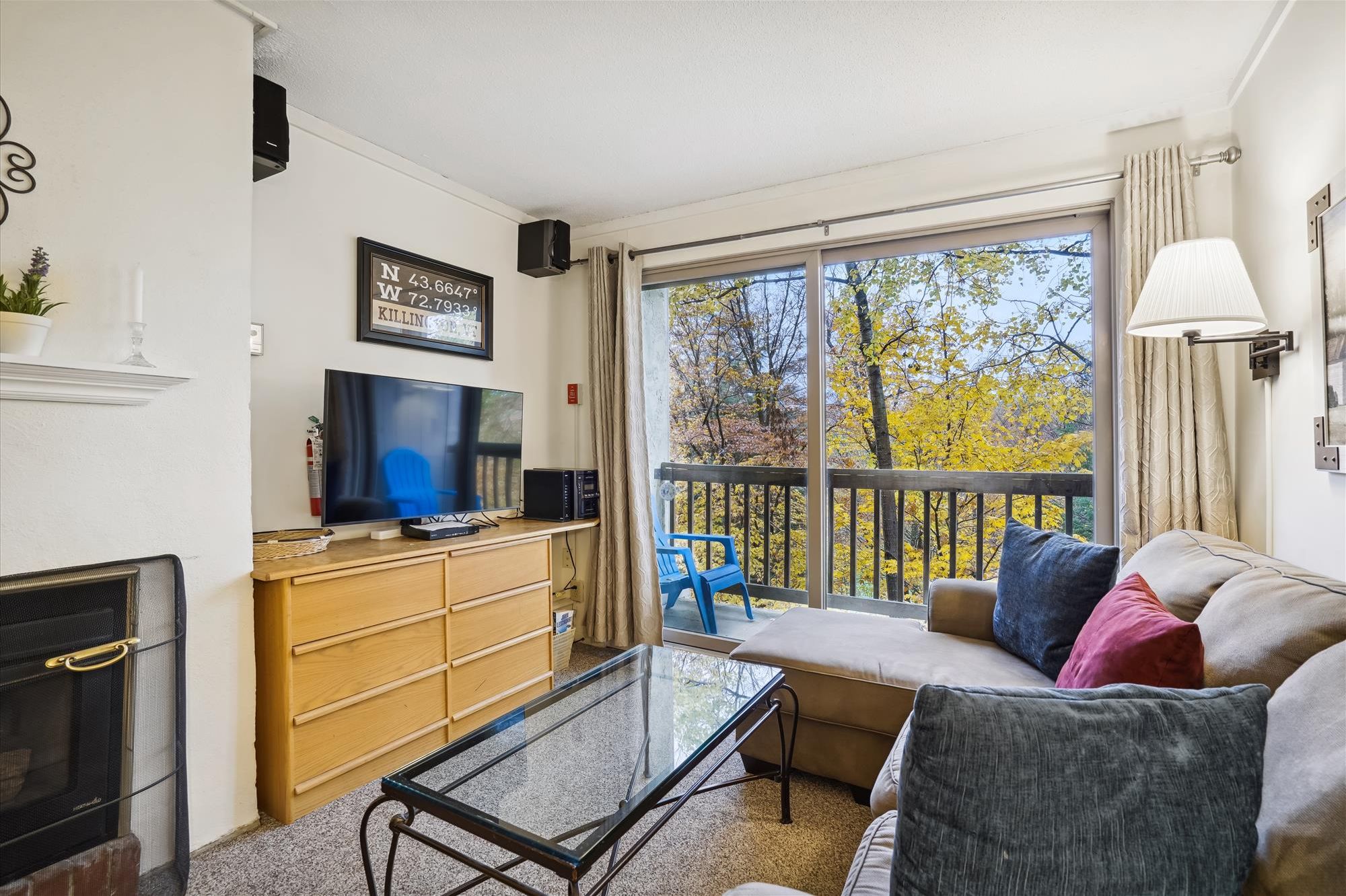 A living room with a TV, wooden cabinets, a glass coffee table, a sofa with cushions, a fireplace, and a balcony with autumn trees.