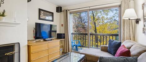 A living room with a TV, wooden cabinets, a glass coffee table, a sofa with cushions, a fireplace, and a balcony with autumn trees.