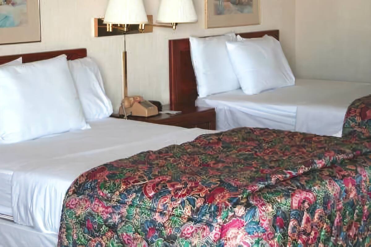 A hotel room with two beds, each with white linens and a colorful floral bedspread. There are two wall-mounted lamps and framed pictures above the beds.