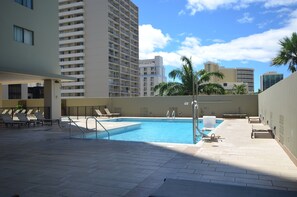 A rooftop pool area with a clear sky, palm trees, and high-rise buildings in the background.