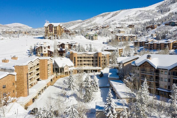 A snow-covered resort with multiple buildings, including a large hotel with a ski lift visible in the background.