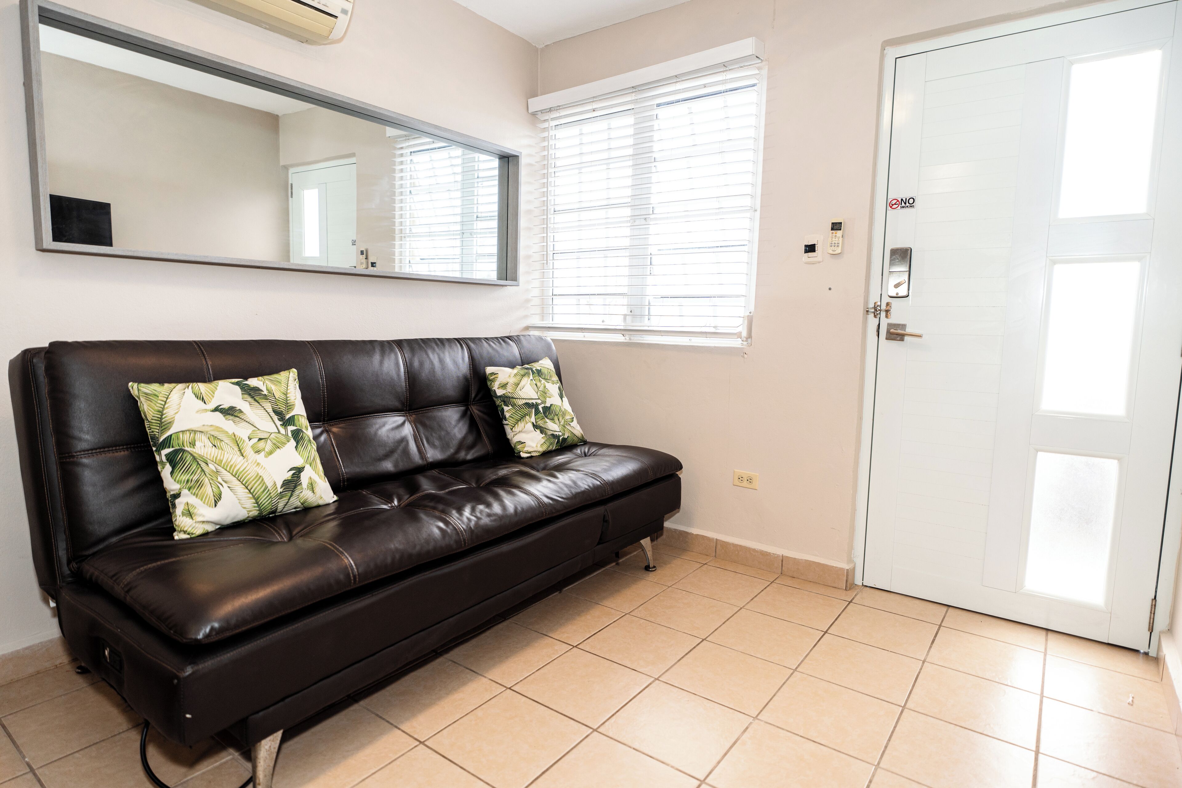 Una habitación con un sofá de cuero negro, dos almohadas con estampado de hojas, un espejo grande, una puerta blanca y una ventana con persianas.