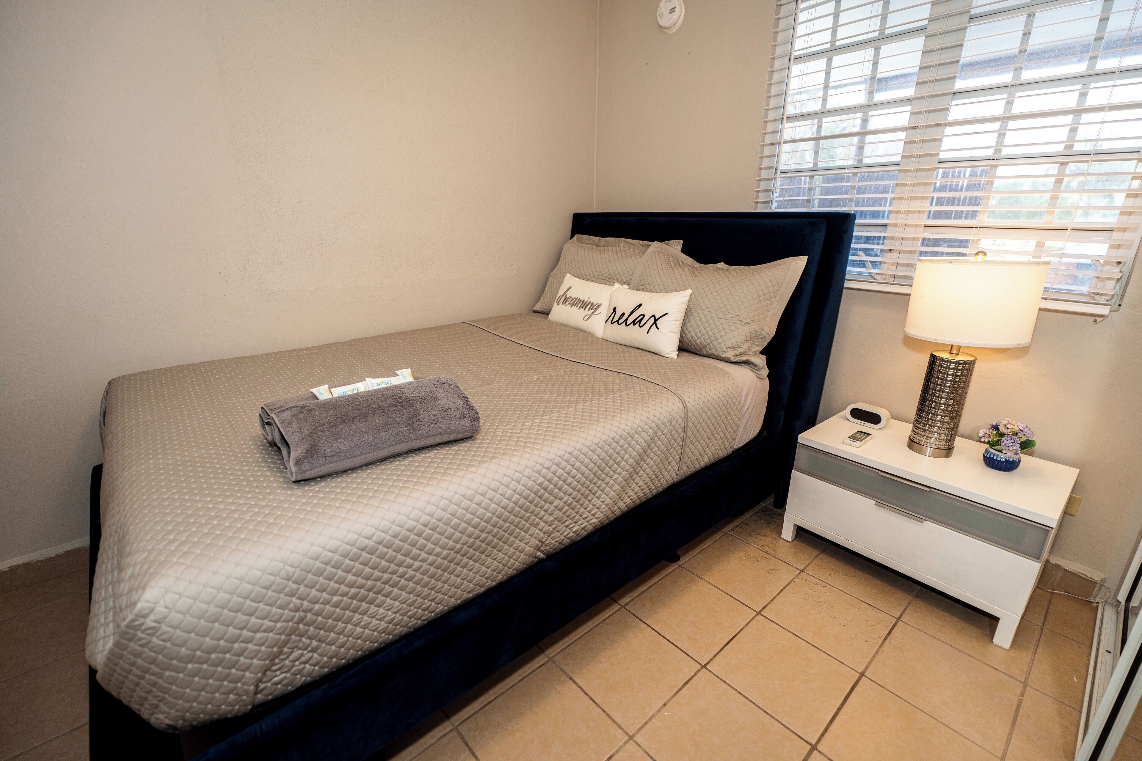 A neatly made bed with a quilted cover, a pillow with the text "Enjoying relax," a folded towel, and a bedside lamp on a white nightstand.