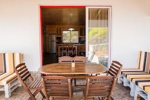 Un coin repas avec une table ronde en bois et des chaises, une cuisine en arrière-plan et une vue sur la verdure à travers une porte vitrée.