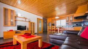 A modern living room with a wooden ceiling, a TV, a dining area with chairs, and a kitchen area with cabinets and a microwave.