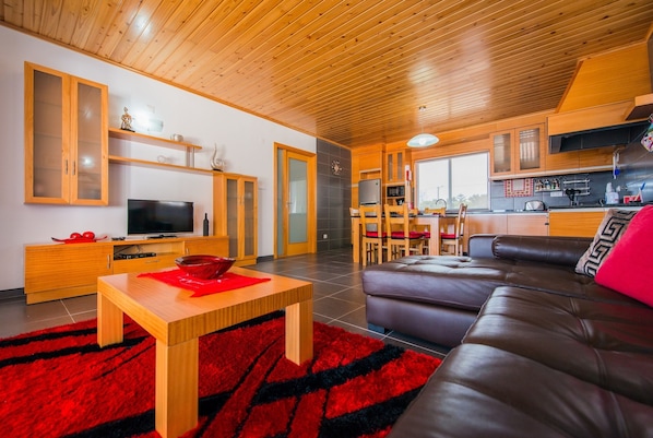 Un salon moderne avec un plafond en bois, un téléviseur, un coin repas avec des chaises et un coin cuisine équipé de placards et d’un four à micro-ondes.