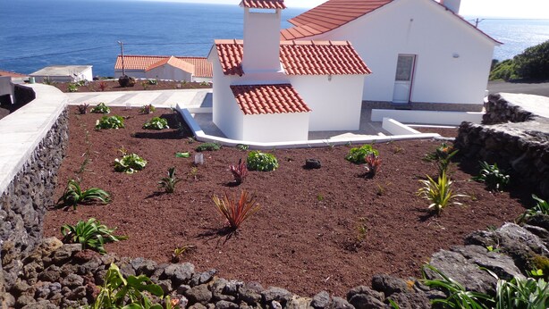 A coastal house with a red-tiled roof, surrounded by a garden with various plants and a rocky border.
