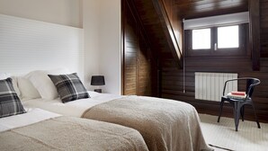 Ein Schlafzimmer mit schräg verlaufender Decke, Holzwänden, einem Bett mit weißer Bettwäsche, einem Sessel mit Buch und einem Fenster mit Jalousien.