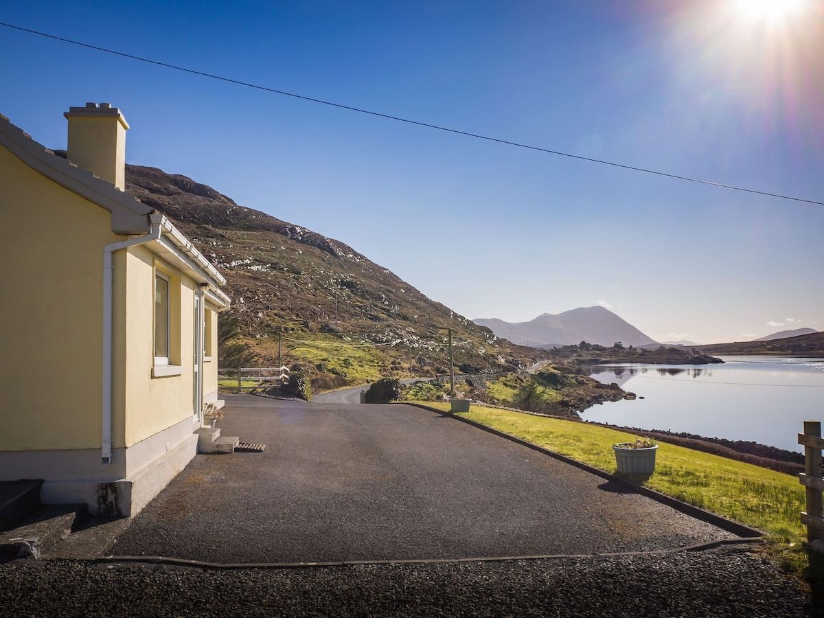 A house with a paved driveway leading to a body of water, surrounded by hills and a clear sky.