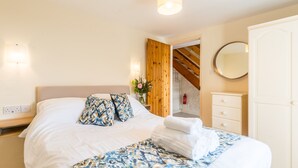 A neatly made bed with a patterned bedspread, a bedside table with a vase of flowers, and a wooden door leading to another room. - 4* self catering, farm stay cottage. Sleeps 4 (Boscastle)