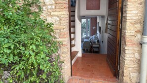 Un muro de piedra con un aro metálico, una puerta de madera y una escalera que conduce a un dormitorio con una cama, un escritorio y una ventana.