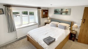 Een slaapkamer met een groot bed, een raam met gordijnen, een nachtkastje met een lamp en een schilderij aan de muur.