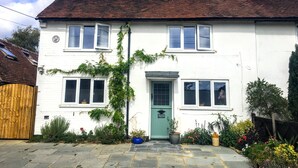 Een wit, twee verdiepingen tellend huis met een groene deur, meerdere ramen, een schoorsteen en een tuin met verschillende planten en bloemen.
