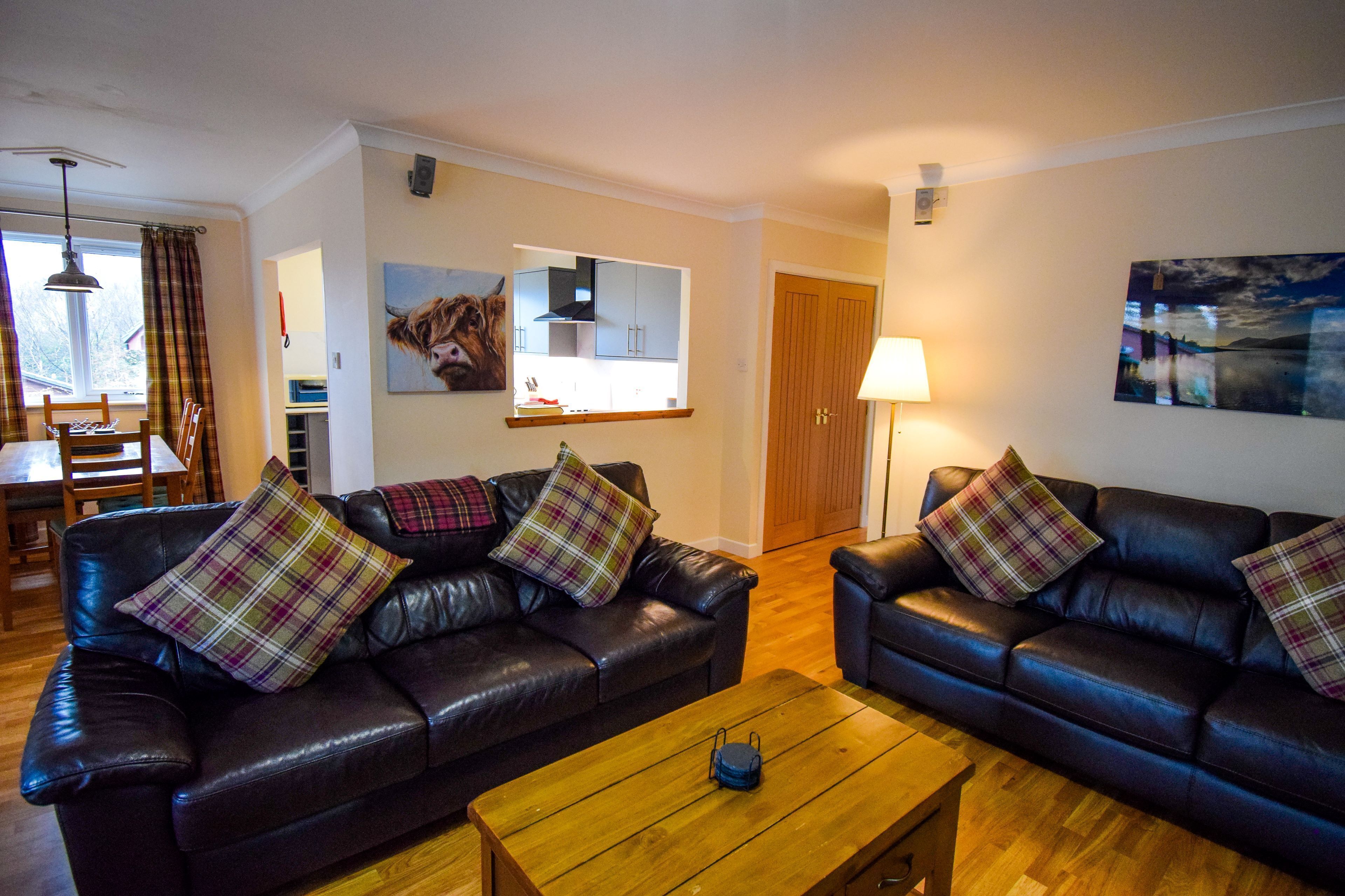 Ein Wohnzimmer mit zwei Ledersofas, einem Holztisch und karierten Kissen. Es gibt einen Essbereich mit Tisch und Stühlen, und durch eine offene Tür ist eine Küche zu sehen.