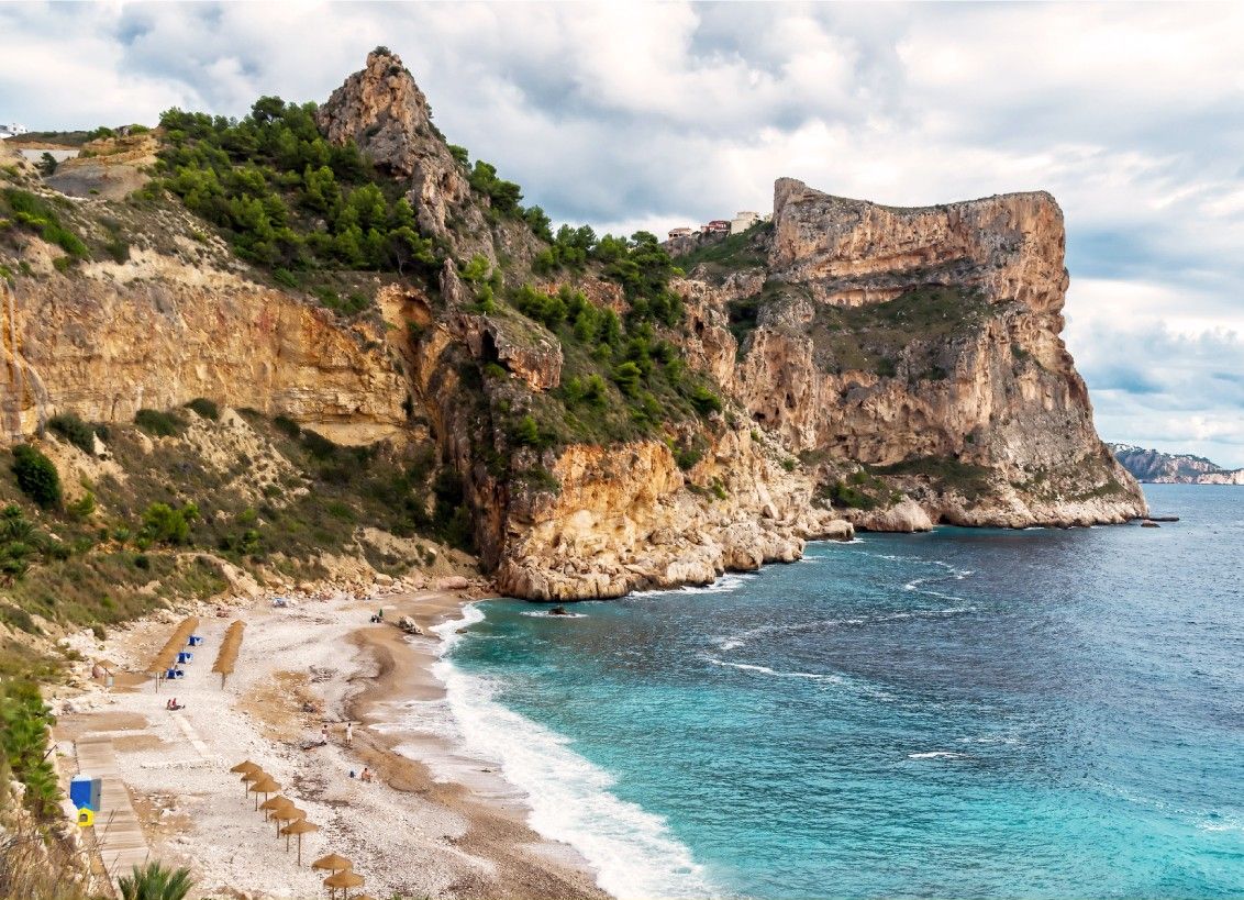 A coastal landscape with a prominent rocky cliff and a beach with umbrellas.