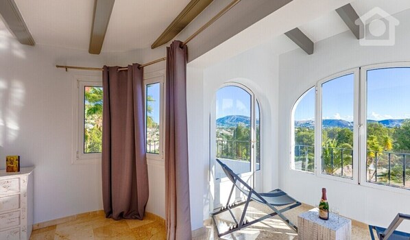 A balcony with a view of mountains and greenery, a chair, and a bottle of champagne.