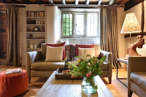 A cozy living room with a sofa, a coffee table with a vase of flowers, a bookshelf, and a window with curtains.