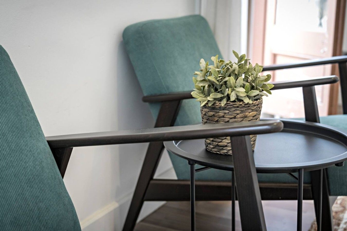 A chair with a woven plant pot on a small table, a teal upholstered chair, and a white wall.