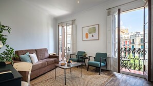 A modern living room with a brown sofa, a coffee table, and two green armchairs. A large window offers a view of the city. - Barcelona Balconies 4 - Habitat Apartments (Barcelona)
