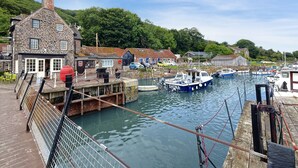 A stone building with a dock extending into a calm harbor, surrounded by greenery and other buildings.