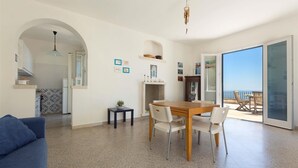 Un vaste espace de vie comprenant une table à manger en bois, des chaises blanches, un canapé bleu et un balcon offrant une vue sur la mer.