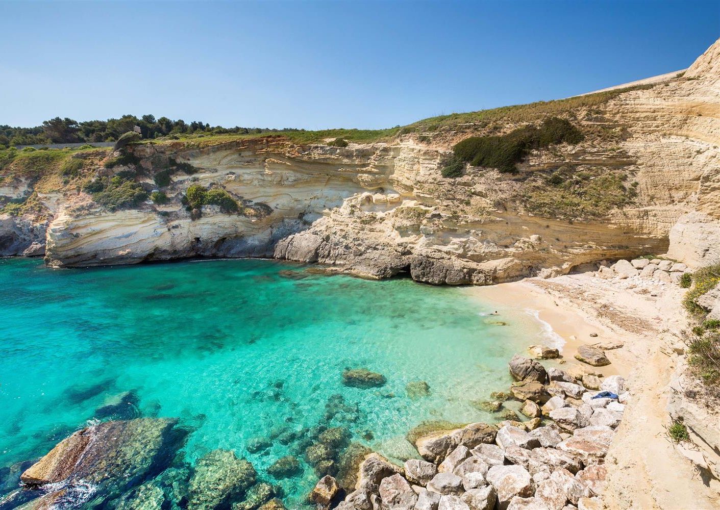 Eine Küstenlandschaft mit klarem türkisblauem Wasser, felsigem Gelände und einem Feldweg, der zu einem Strand führt.