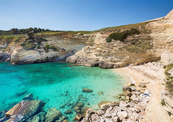 A coastal landscape with clear turquoise waters, rocky terrain, and a dirt path leading to a beach.