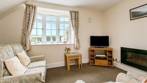 A cozy living room with a sofa, a small table with a book and vase, a TV on a wooden stand, and a fireplace. - Four star two bedroom cottage both with en suite facilities (Minehead)