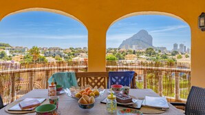 Un balcón con una mesa dispuesta para el desayuno, vista a una montaña y un paisaje urbano al fondo.