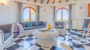 Una habitación con suelo a cuadros, muebles de mimbre, una mesa redonda de cristal y dos ventanas arqueadas con cortinas.