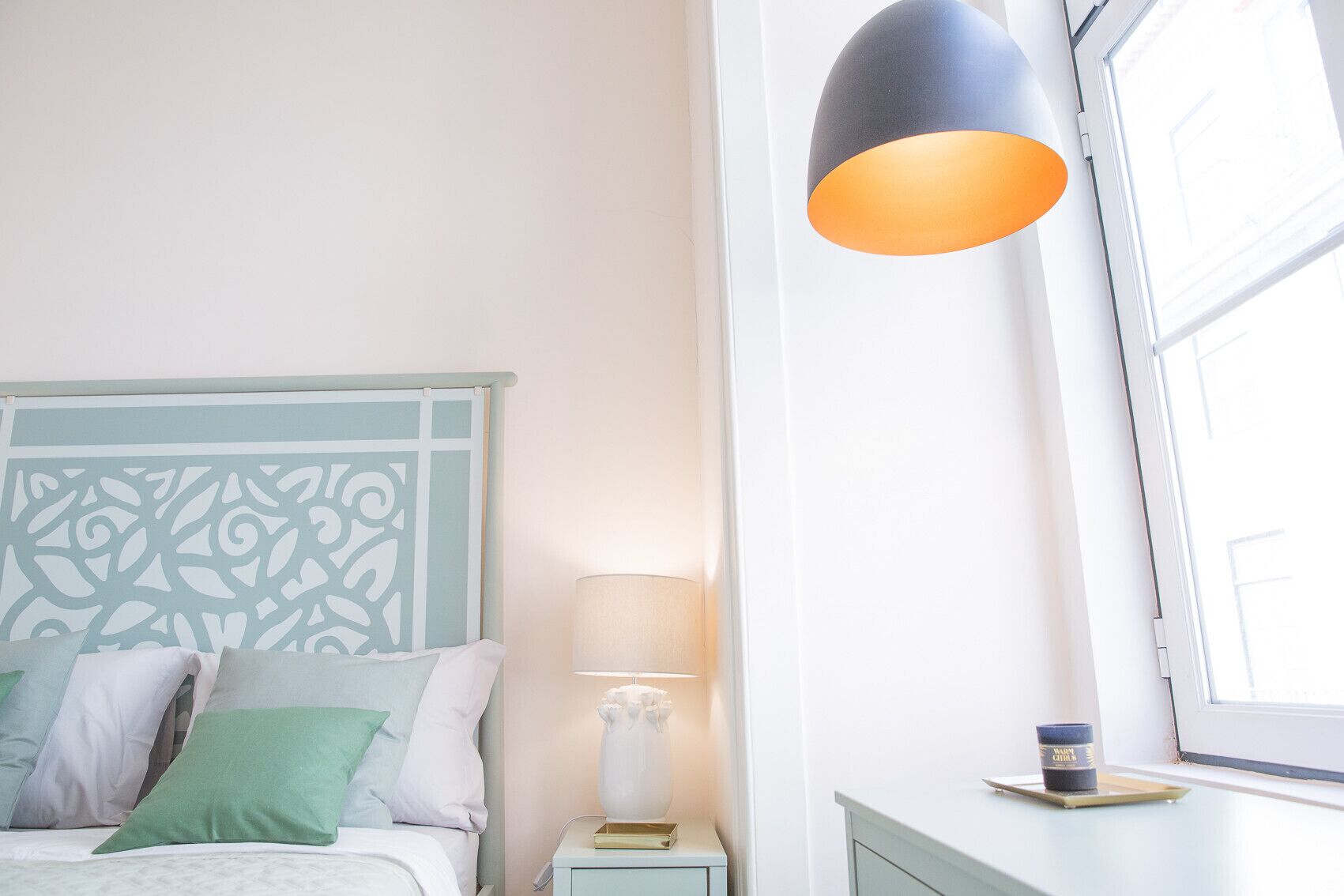 A bedroom with a bed, a nightstand, a lamp, a window, and a view of the outside.