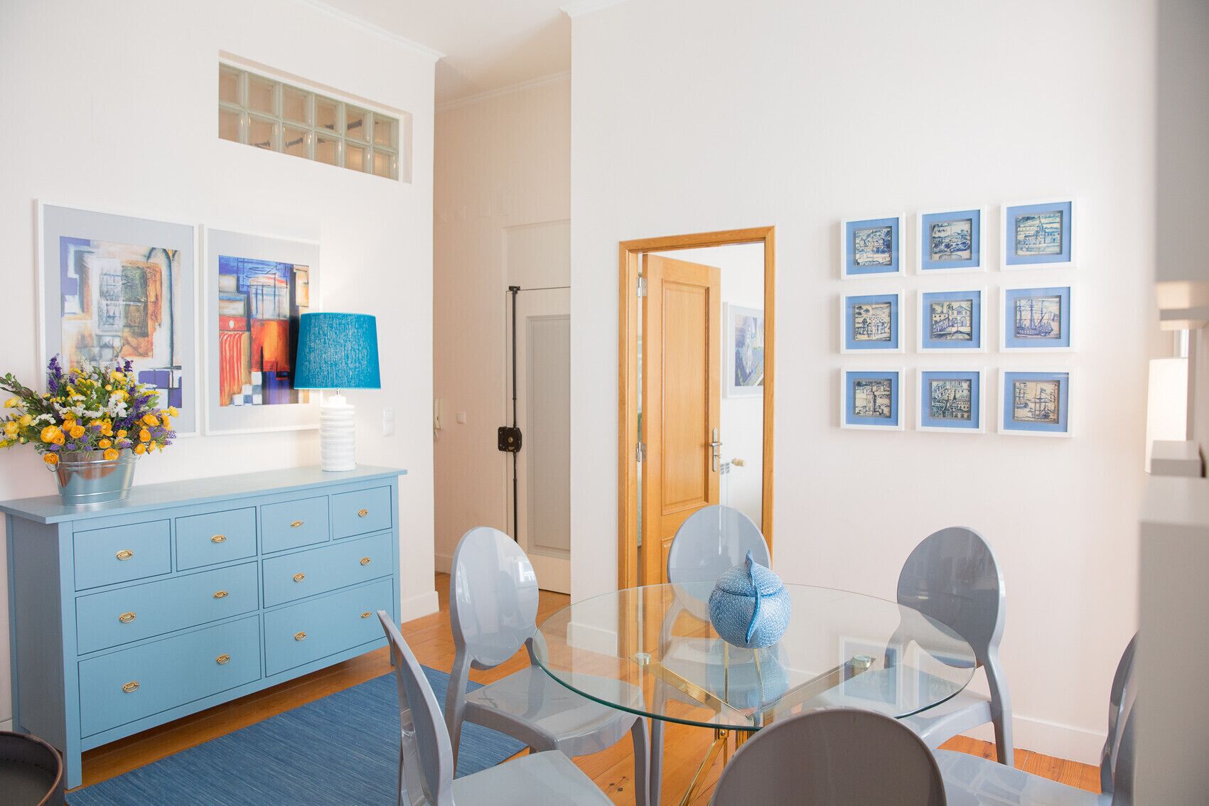 A dining area with a glass table and chairs, a blue cabinet with drawers, and framed artwork on the walls.