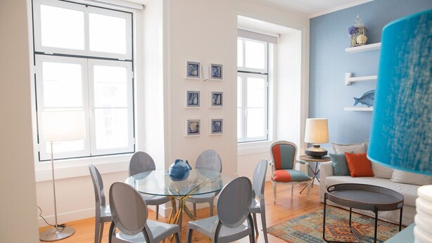 Een moderne woonkamer met een glazen eettafel, grijze stoelen, een blauwe bank en een kamerbreed tapijt met een patroon.