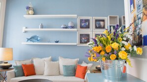 Een woonkamer met een bank, een bijzettafeltje en een bloevas. Er zijn planken met decoratieve objecten en ingekaderde foto's aan de muur.