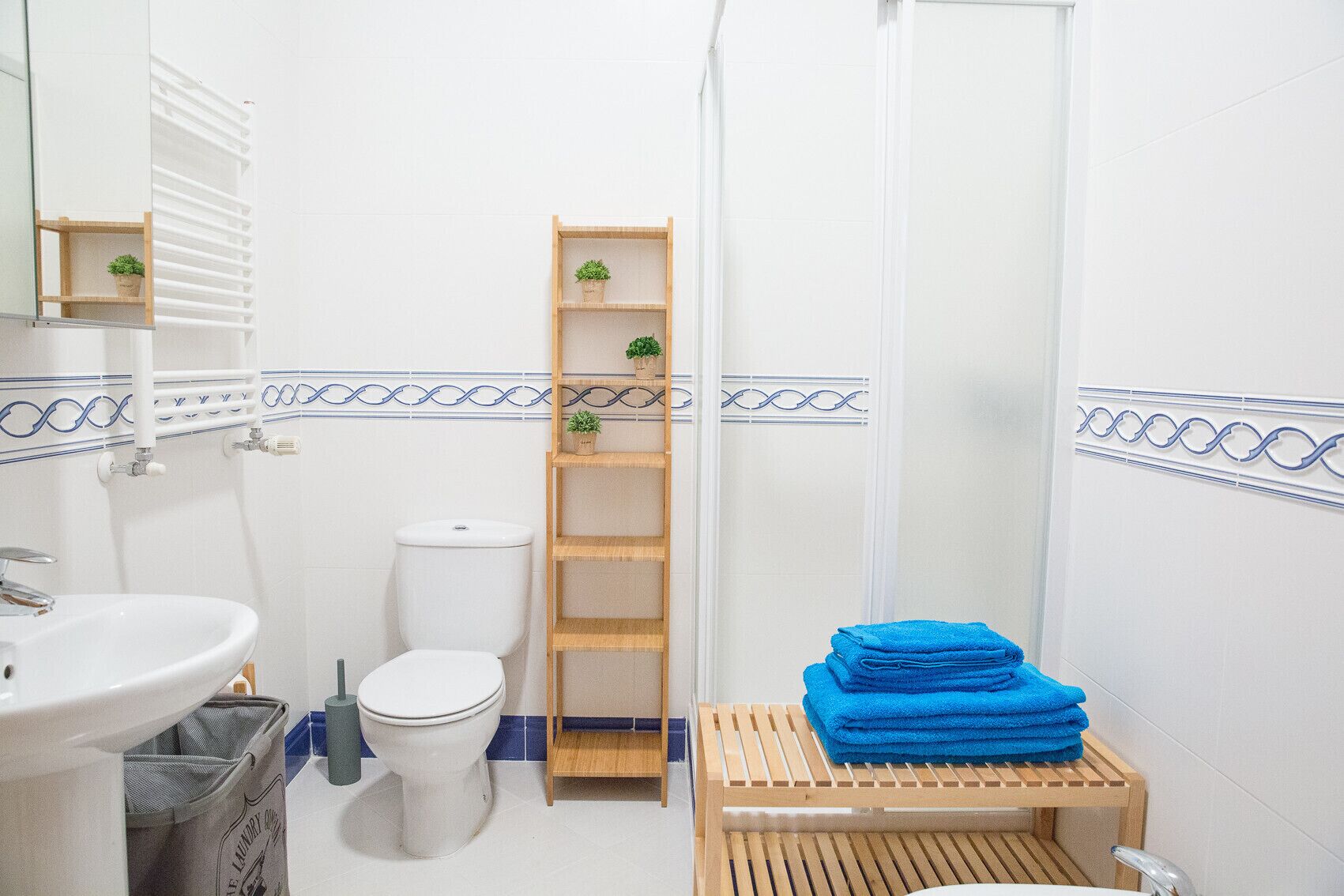 A bathroom with a white sink, toilet, and shower area. There is a wooden shelf with plants and a stack of blue towels on a wooden rack.