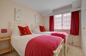 A bedroom with two beds, each with a white headboard and red bedding. There is a window with red curtains, a bedside table with a lamp, and floral artwork on the wall. - 2 Bed in Instow (oc-n1093) (Instow)