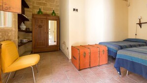 A room with a wooden wardrobe, a yellow chair, two beds with blue bedding, and a suitcase on the floor.