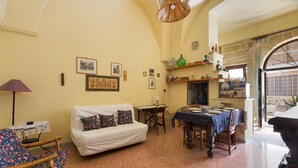 A cozy living room with a sofa, dining table, and chairs. There are framed pictures on the wall and a window with a view.
