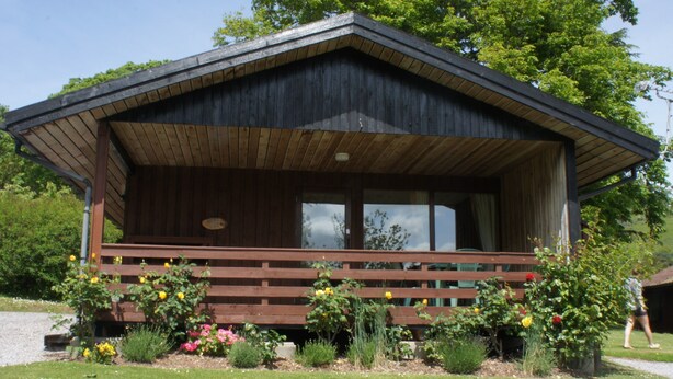 A wooden cabin with a porch, surrounded by a garden with various plants and flowers. - Four star Lodge with two bedrooms, fabulous views yet close to pubs and beach (Minehead)
