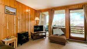 A cozy room with wooden walls, a television on a wooden stand, a sofa with cushions, and a view through a glass door to an outdoor deck. - Four star Lodge with two bedrooms, fabulous views yet close to pubs and beach (Minehead)