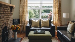 A living room with a fireplace, a sofa, an ottoman, and a large window with curtains. - Portrush - Hope Cottage - sleeps 5 guests  in 3 bedrooms (Portrush)