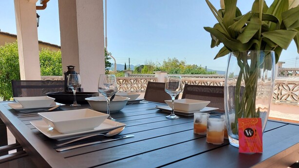 Una mesa lista para una comida, con platos, tazones, vasos y un jarrón con hojas verdes.