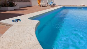 Una piscina con agua cristalina azul, rodeada por un muro blanco y un pilar de piedra. Hay dos sillas y una mesa cerca de la piscina.