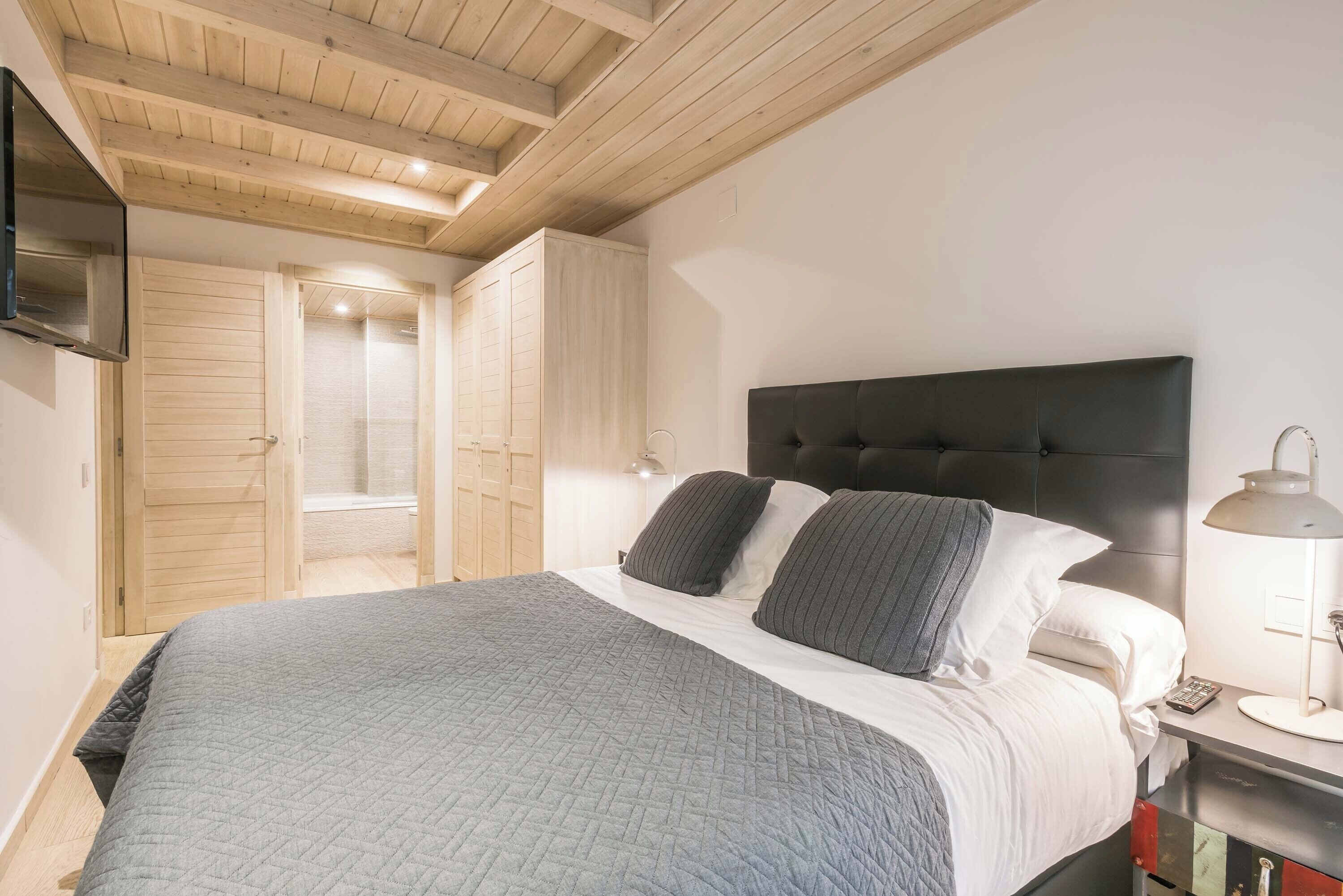 A bedroom with a bed, a nightstand, a TV mounted on the wall, and wooden ceiling beams.