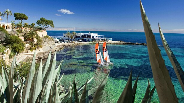 Un hotel sulla costa con acque cristalline, palme e una barca a vela.